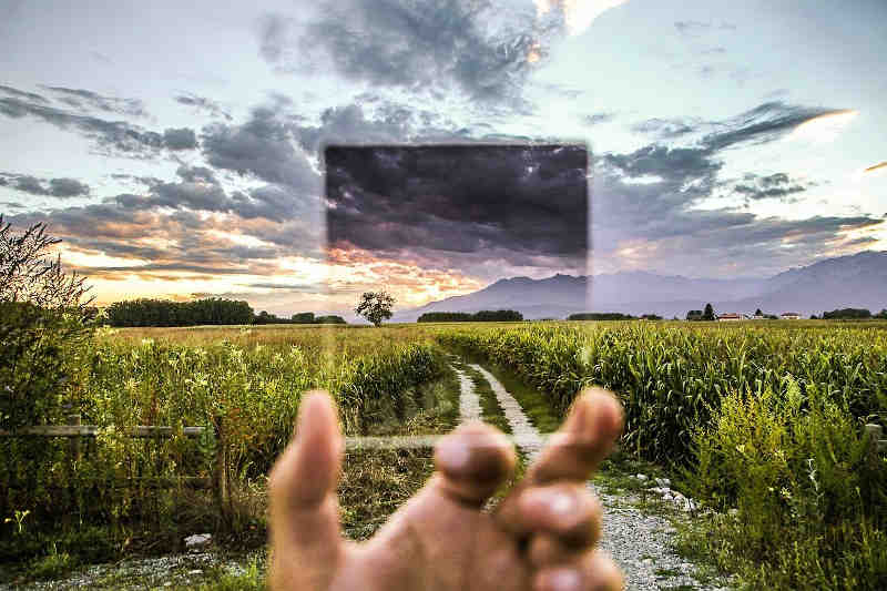 Country landscape with central track leading-away through a field, distanced hills, and dramatic sky with grey clouds and sunset colours; partially seen through a hand-held square of glass that's a darkening filter.